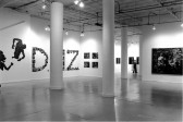 A photograph of a large gallery space with white walls and columns in the center of the room and artworks lining the walls.  In the center of the image, visible through the columns, are many small photographs hung in the shape of the letters 