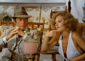 A man and a woman sit in two low wooden chairs. The man wears a brown hat and striped suit, while the woman wears a white top with a low v-neck. Her hair is blonde and stylishly curled. She stares ahead, her left hand gently resting on her chin. The man stares sideways at her.