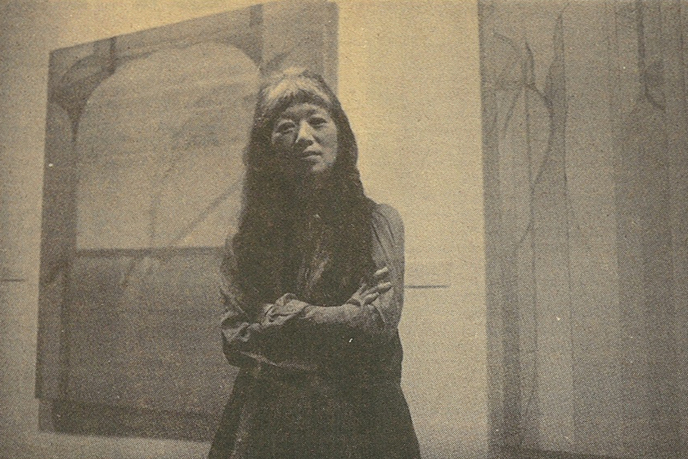 Faded photograph of artist Miyoko Ito standing with her arms crossed in front of two of her paintings.