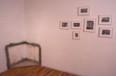 The corner of a gallery space. At the base of the corner, where both walls meet the floor, a small area of the wall has been removed and replaced with the materials from another building's wall, displayed in a cross-section. Several black and white, framed photo-collages hang on the wall to the right of the corner.