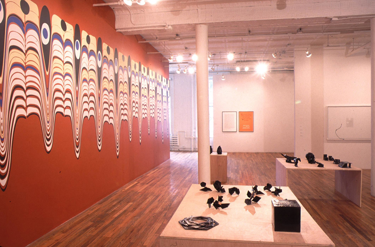 Several small, model-like black sculptural forms displayed on low wooden supports in the center of a large gallery space with wooden flooring. A large wall decal featuring eye-like forms and swirling, dripping stripes of color covers the wall to the left of the sculptures. Additionally, several framed 2D works hang on the back gallery walls behind the displayed sculptures.
