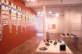 Several small, model-like black sculptural forms displayed on low wooden supports in the center of a large gallery space with wooden flooring. A large wall decal featuring eye-like forms and swirling, dripping stripes of color covers the wall to the left of the sculptures. Additionally, several framed 2D works hang on the back gallery walls behind the displayed sculptures.