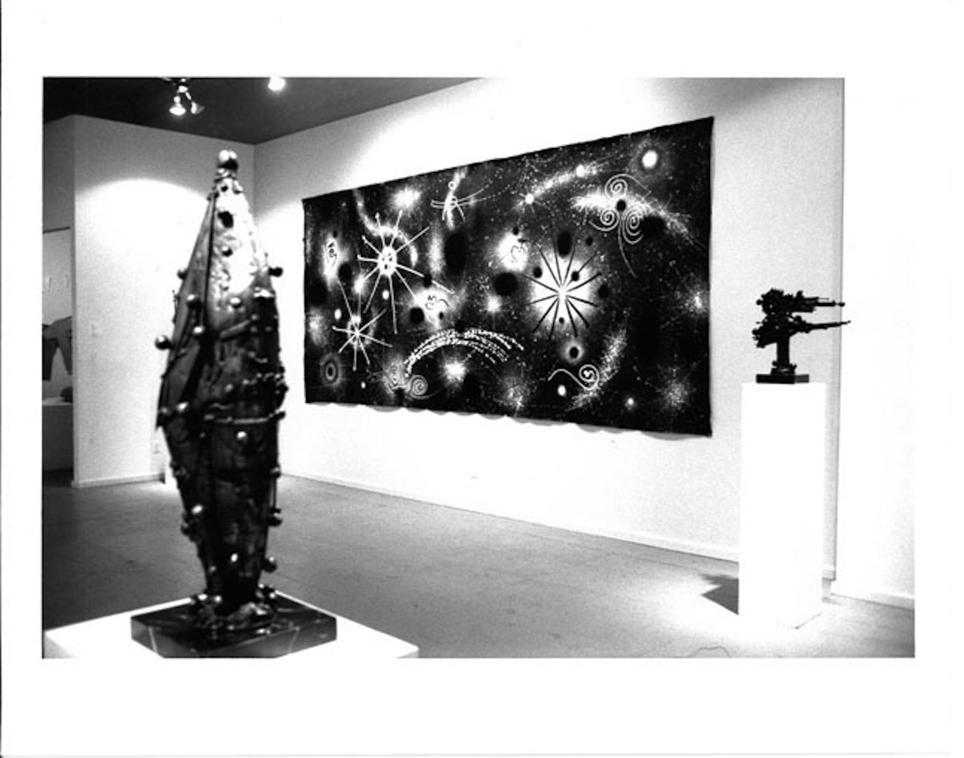 Two sculptures appear on pedestals in the foreground and a painting is visible in the background in a white-walled gallery space.