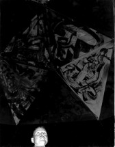 A black and white photograph of a painted, sculpted object appears hanging above a person's head in a dark space.