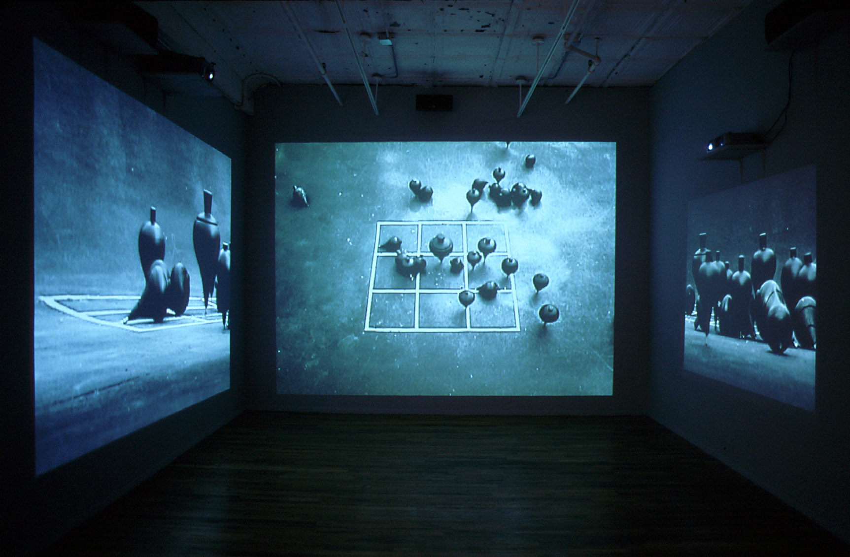 A small, empty room with images projected on three walls. All are gray/blue shots of spinning tops on a white grid on concrete. The center image shows this from above, while the left and right images show the tops from the side.