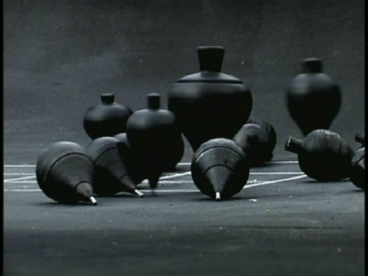 A collection of black spinning tops on concrete. Several are upright, spinning, and several have fallen onto their sides. A white grid is just visible on the ground beneath them.