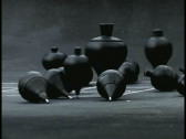 A collection of black spinning tops on concrete. Several are upright, spinning, and several have fallen onto their sides. A white grid is just visible on the ground beneath them.