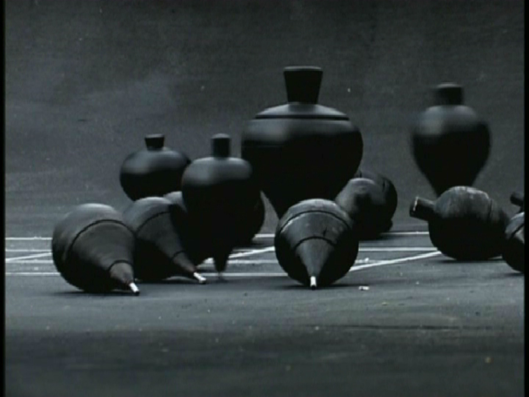 A collection of black spinning tops on concrete. Several are upright, spinning, and several have fallen onto their sides. A white grid is just visible on the ground beneath them.