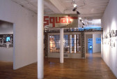 A gallery space with two white columns bisecting the room. There are black and white images on the wall and a large sign suspended from the ceiling with red letters reading 
