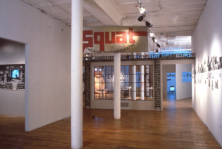 A gallery space with two white columns bisecting the room. There are black and white images on the wall and a large sign suspended from the ceiling with red letters reading 