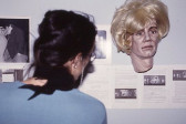 A person looks at several sheets of paper with text and black and white photographs mounted on a wall. Hanging just above the papers is a sculpture of a human head with blonde wig.