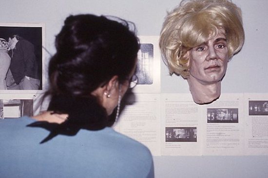 A person looks at several sheets of paper with text and black and white photographs mounted on a wall. Hanging just above the papers is a sculpture of a human head with blonde wig.