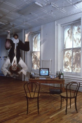 A room with two chairs facing toward a low table with a small monitor and assorted bottles. There are two windows, both blotted out with smears of paint. A large sculpture of a person with crouched legs and their arms above their head is suspended from the ceiling, with a second smaller head emerging from between the squatting legs.