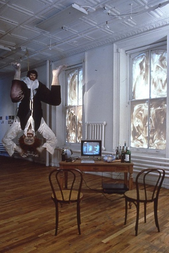 A room with two chairs facing toward a low table with a small monitor and assorted bottles. There are two windows, both blotted out with smears of paint. A large sculpture of a person with crouched legs and their arms above their head is suspended from the ceiling, with a second smaller head emerging from between the squatting legs.