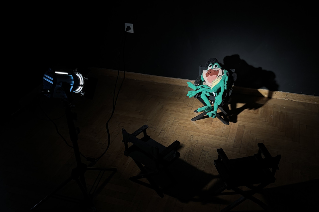 In a dark room with a black wall and wood floor, there is a spotlight shining on two frog plushies. One sits horizontally in the lap of another who is seated in a director's chair. Two more director's chairs, not illuminated, sit facing the frogs. The spotlight, the chairs, and the frogs all appear miniature-sized.