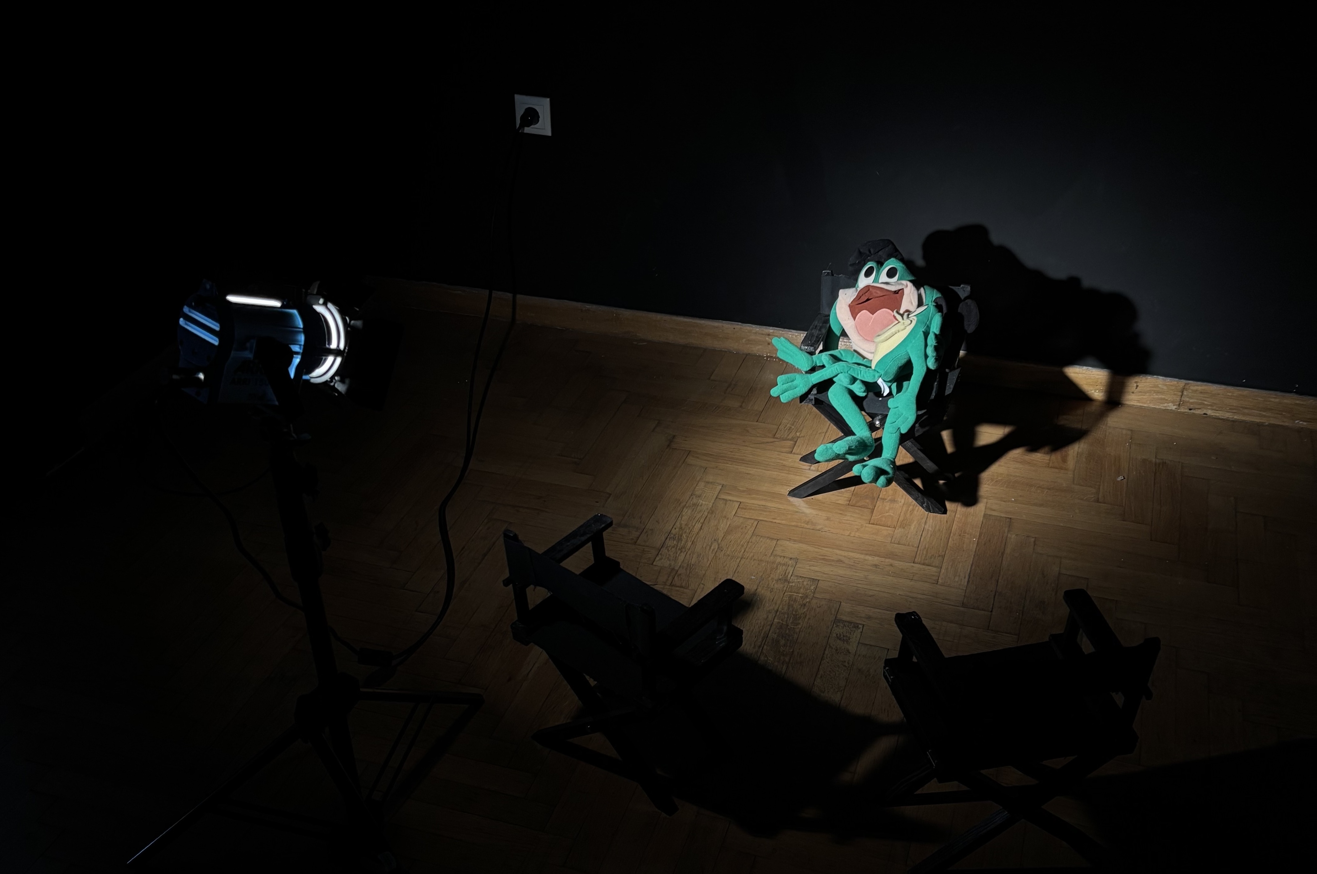 In a dark room with a black wall and wood floor, there is a spotlight shining on two frog plushies. One sits horizontally in the lap of another who is seated in a director's chair. Two more director's chairs, not illuminated, sit facing the frogs. The spotlight, the chairs, and the frogs all appear miniature-sized.