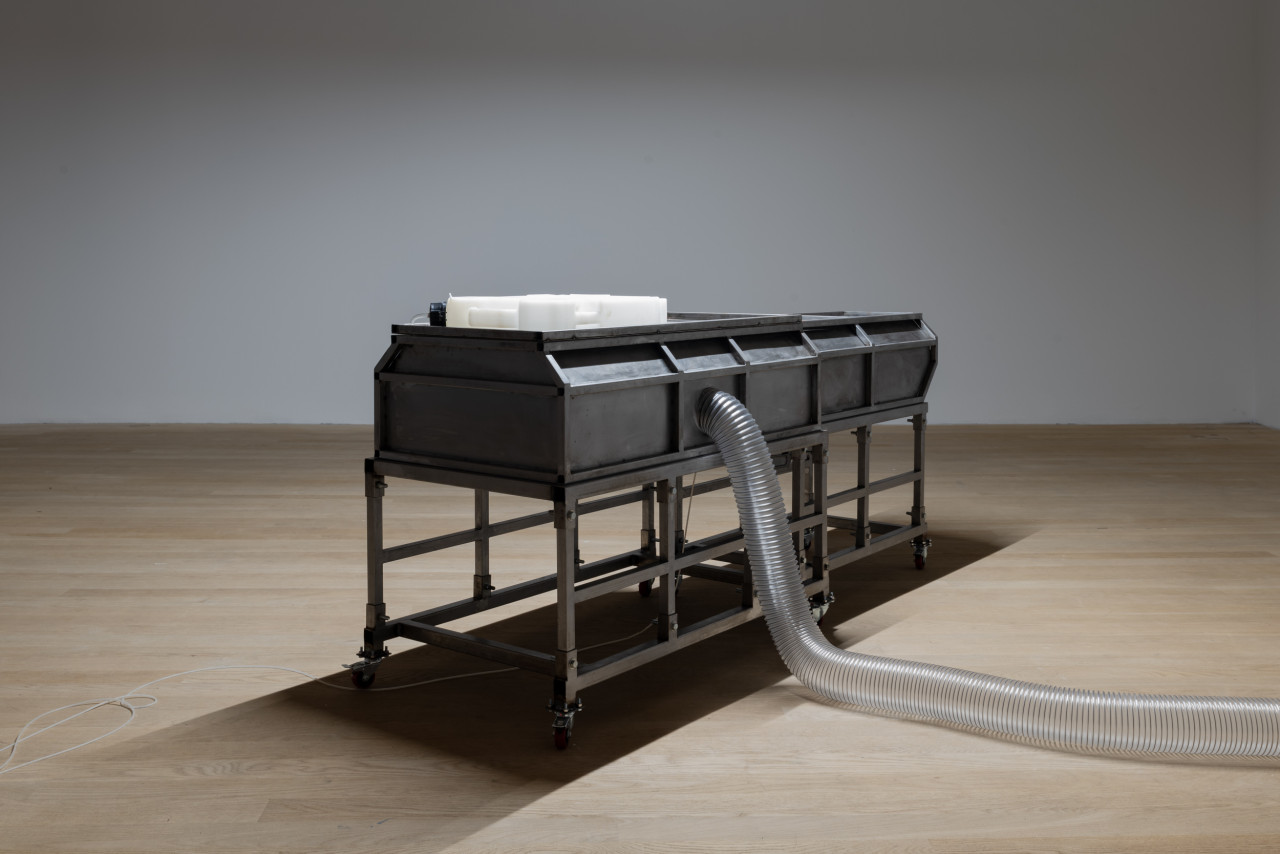 A human-sized steel box stands in the room, lit by white light from above. The box rests on metal supports, and a plastic tube protrudes from one side.