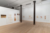 Along two white walls a series of paintings are arranged. Many of the paintings loosely depict human figures or abstracted body parts in brown and red toned paint against white backgrounds.