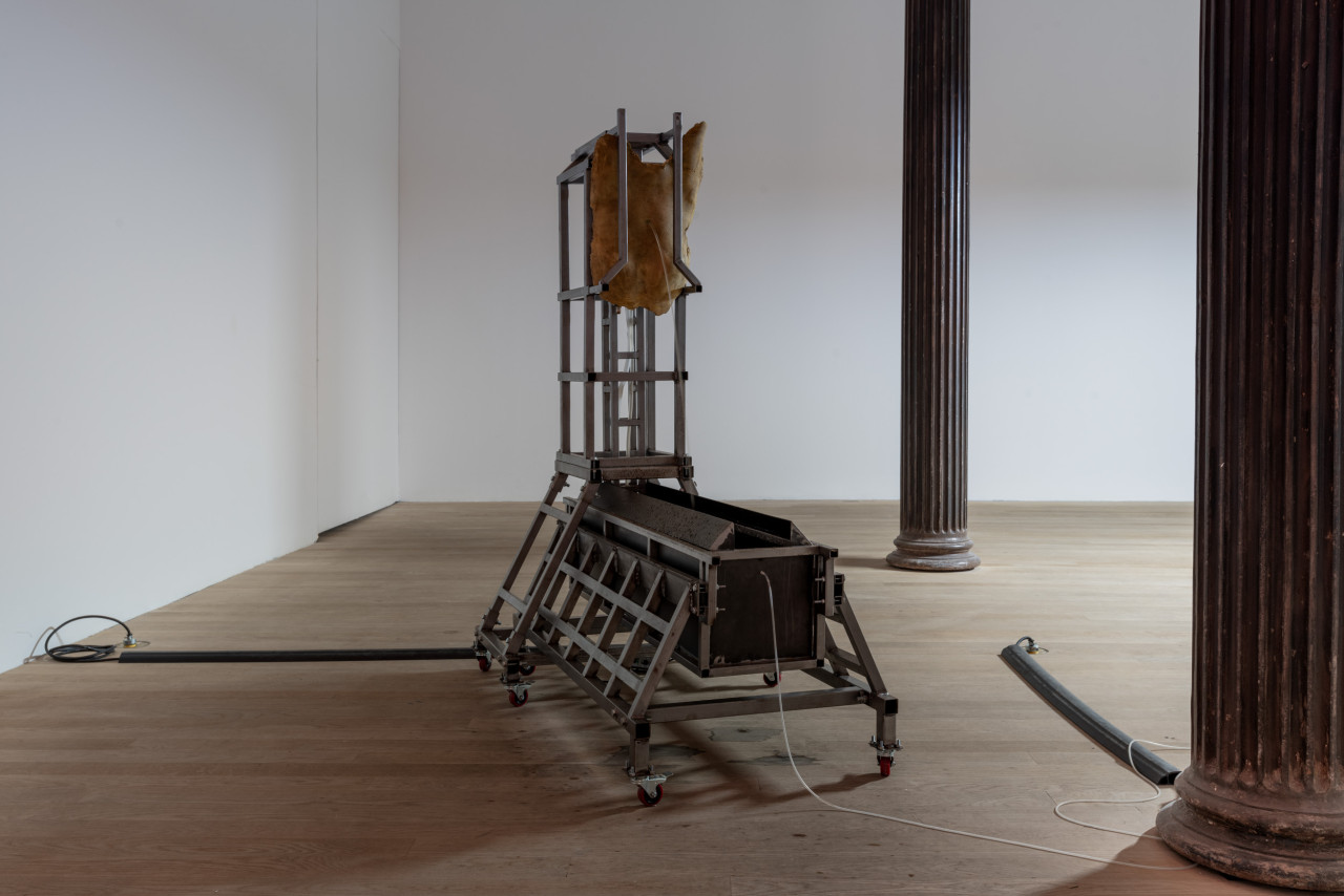 A trough sits between two iron columns, liquid spouts out from the belly button of a torso-like sculpture hanging above the trough.