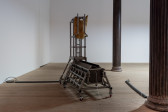 A trough sits between two iron columns, liquid spouts out from the belly button of a torso-like sculpture hanging above the trough.