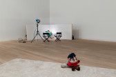 In the foreground a plushie of a cartoon mouse leans on a beaten up Nike Air Force One shoe, and sits on a white carpet. In the background, two panels of white wood, some director's chairs, and a stage light are arranged like a movie set. At the center of the set are two green frog plushies. 