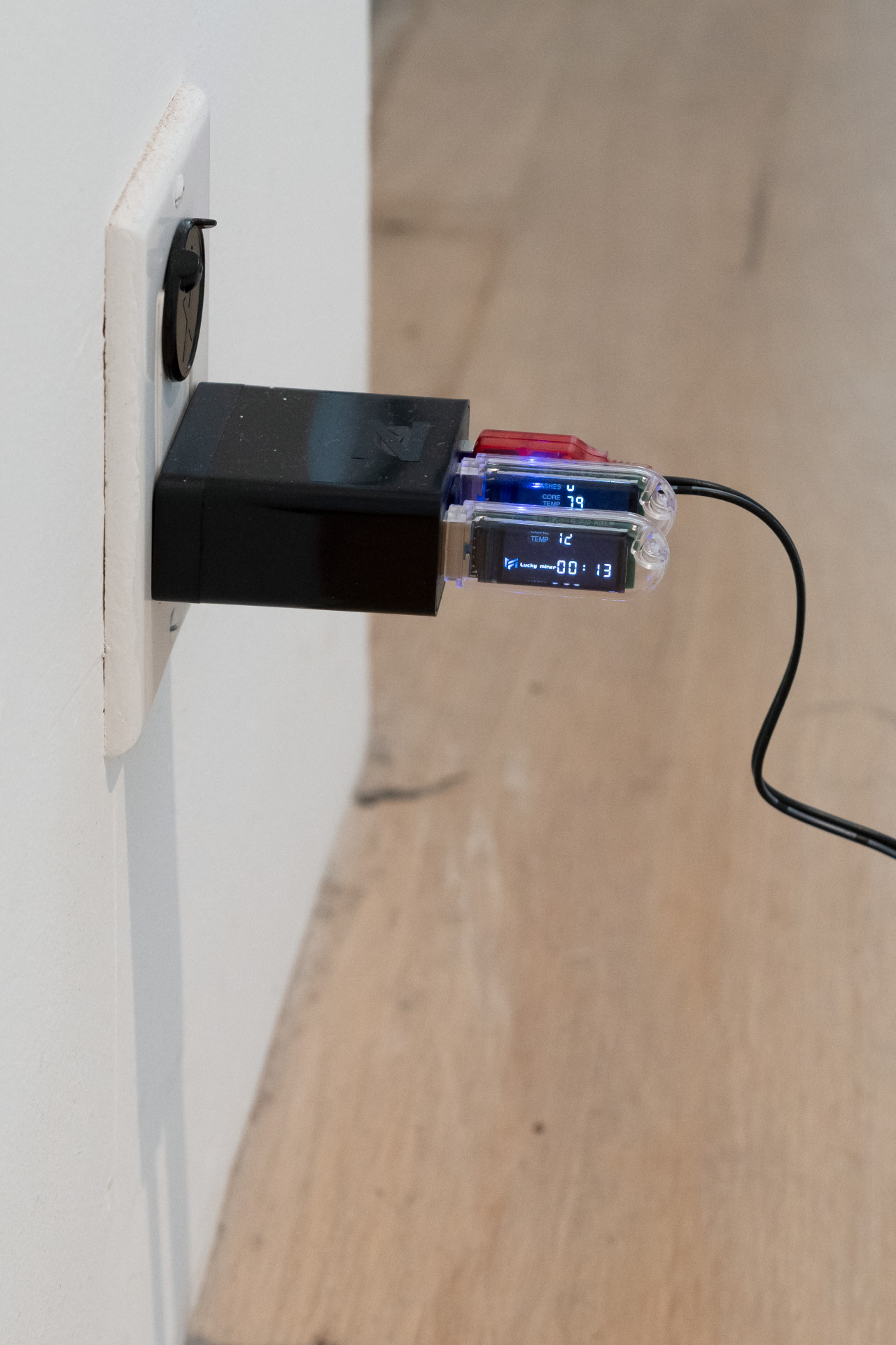 Into an outlet socket on a wall is plugged a large black adapter with two USBs plugged into it. Screens on the USBs flash text that reads "TEMP 12 / Lucky miner 00:13." In the outlet above the USBs is a black plastic cover.