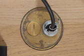 A brass outlet plug against a wooden floor has a cable plugged into one socket, and the other socket is covered by a flat brass circle with the text 
