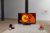 Two toys, sitting on a white rug and propped up by white sneakers, face a TV screen. The TV screen between them displays a scene from the cartoon *Hercules*