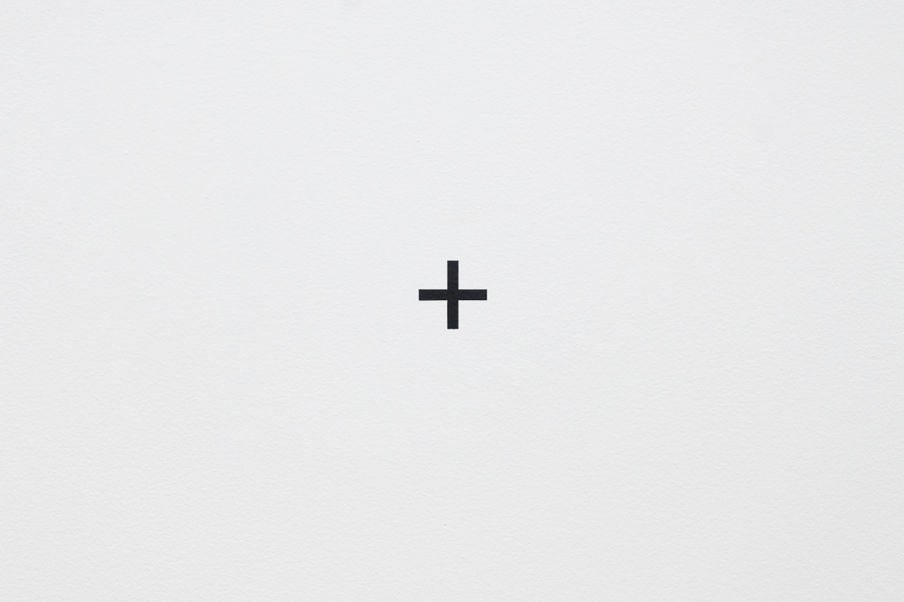 On a white gallery wall, a vinyl cut out of a black cross or "plus-sign" sits perfectly in the center.
