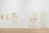 A series of paintings made with brown and red paint against a white background are hung on a wall.