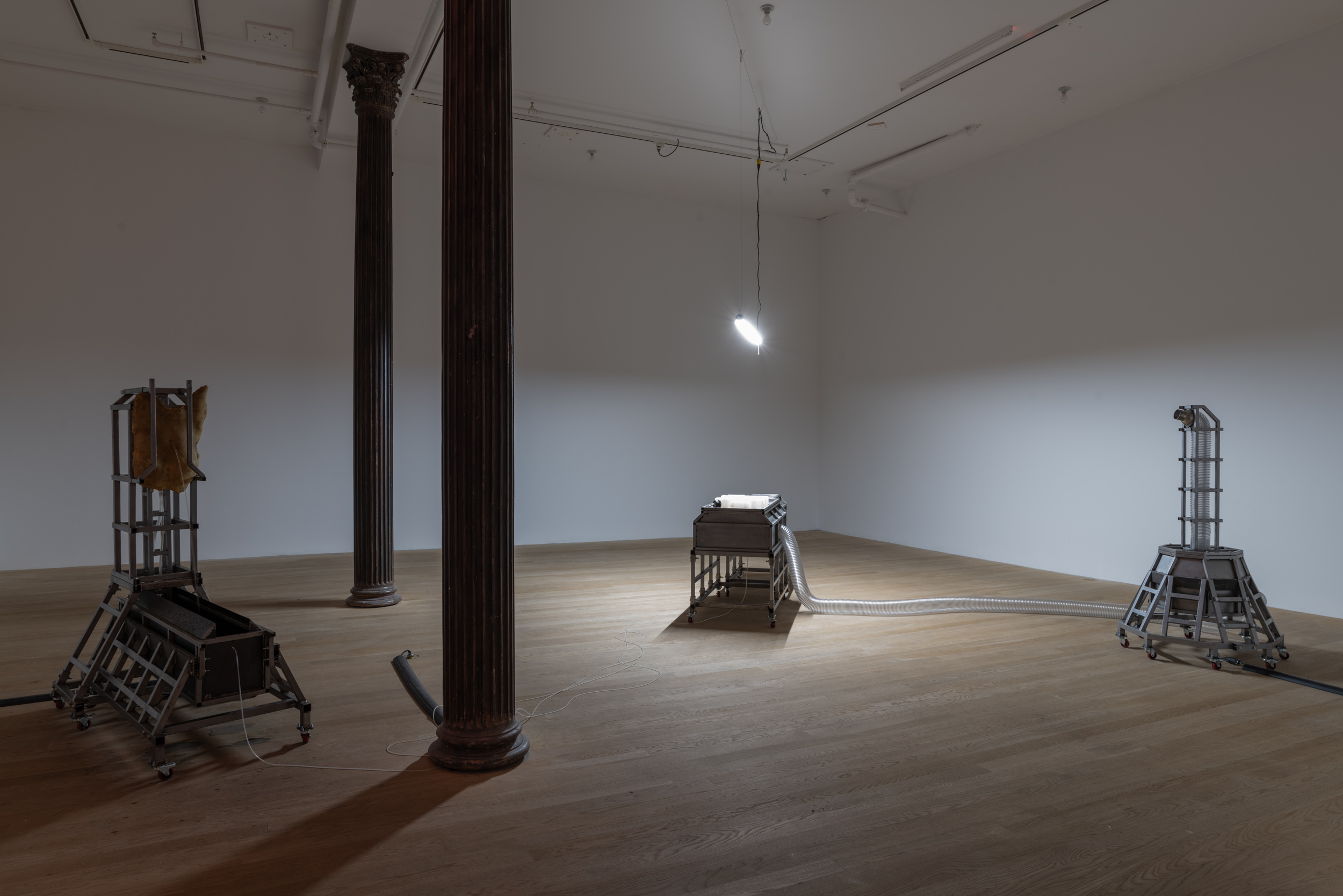 Three structures sit in the space, lit from a singular overhead white LED strip light. On the left sits a trough, fed liquid through the belly button of a torso-like sculpture. In the middle sits a human-sized metal box. A clear tube connects the box to the structure standing on the right, which includes metal rails that hold and pitch the tube upright.