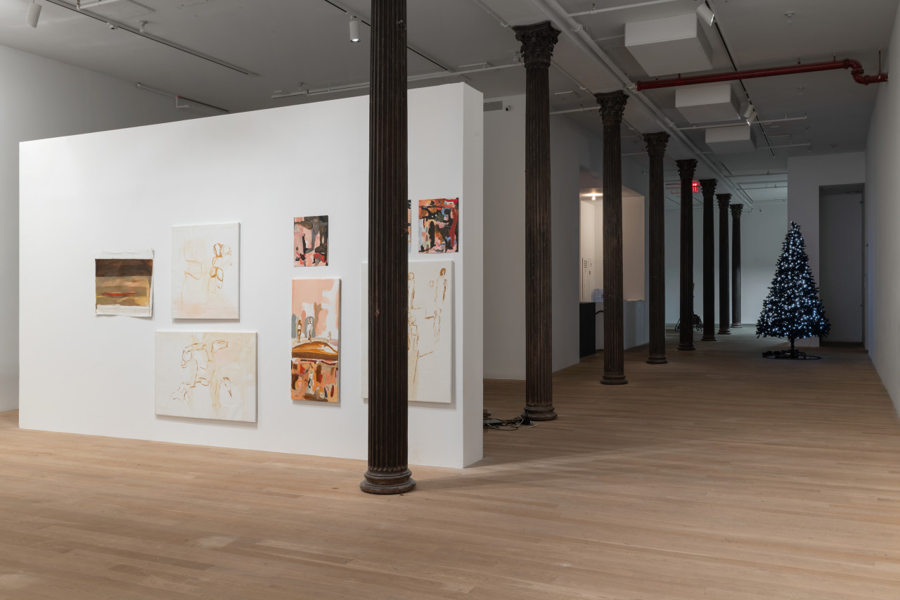 A free-standing wall with paintings on it is placed in the middle of a gallery. Iron columnds run down the center of the gallery, and in the background past the wall is a christmas tree decorated with lights.