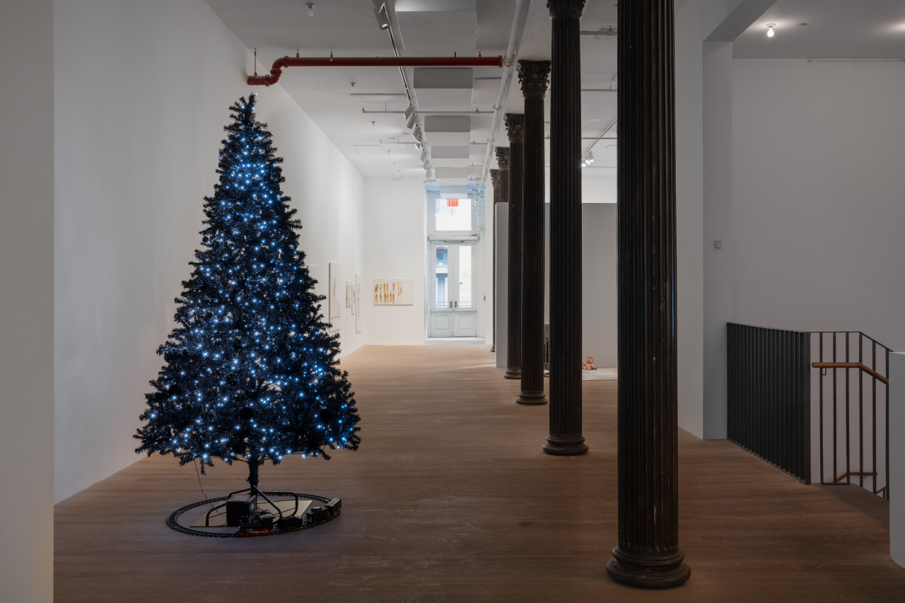 A twinkling black Christmas tree stands in the foreground as the gallery walls recede. A few paintings hang on the more distant walls.