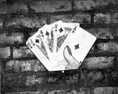 A black and white photograph of a sculptural hand of cards with a ten, jack, queen, and king of diamonds. The piece is placed over a brick surface.