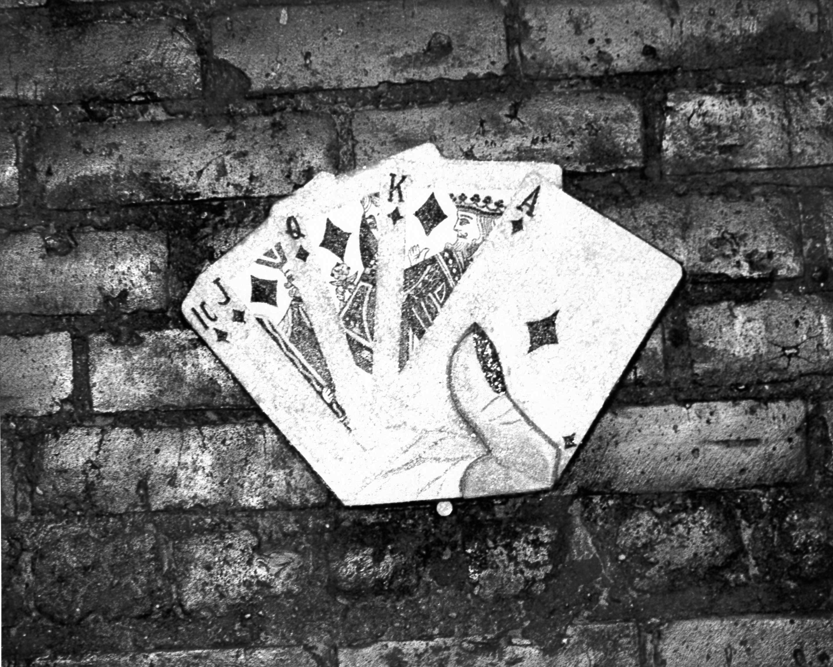 A black and white photograph of a sculptural hand of cards with a ten, jack, queen, and king of diamonds. The piece is placed over a brick surface.