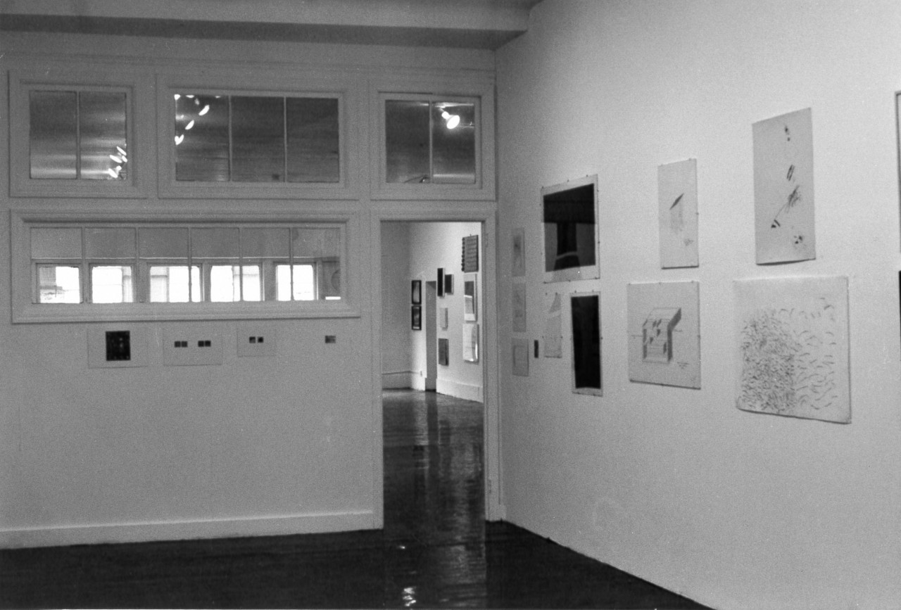 A black and white installation view of a gallery space with windows and a doorframe. Several artworks hang from the walls.