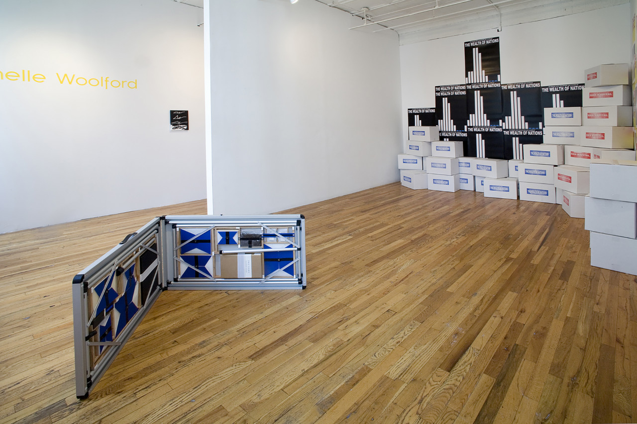 In the foreground is a low sculpture made of folding metal framework that contains blue and white geometric prints as well as boxes. In the background are stacks of boxes that are printed with the words "Das Kapital." Behind them are black and white posters with identical graphs that are titled "The Wealth of Nations."