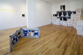 In the foreground is a low sculpture made of folding metal framework that contains blue and white geometric prints as well as boxes. In the background are stacks of boxes that are printed with the words 
