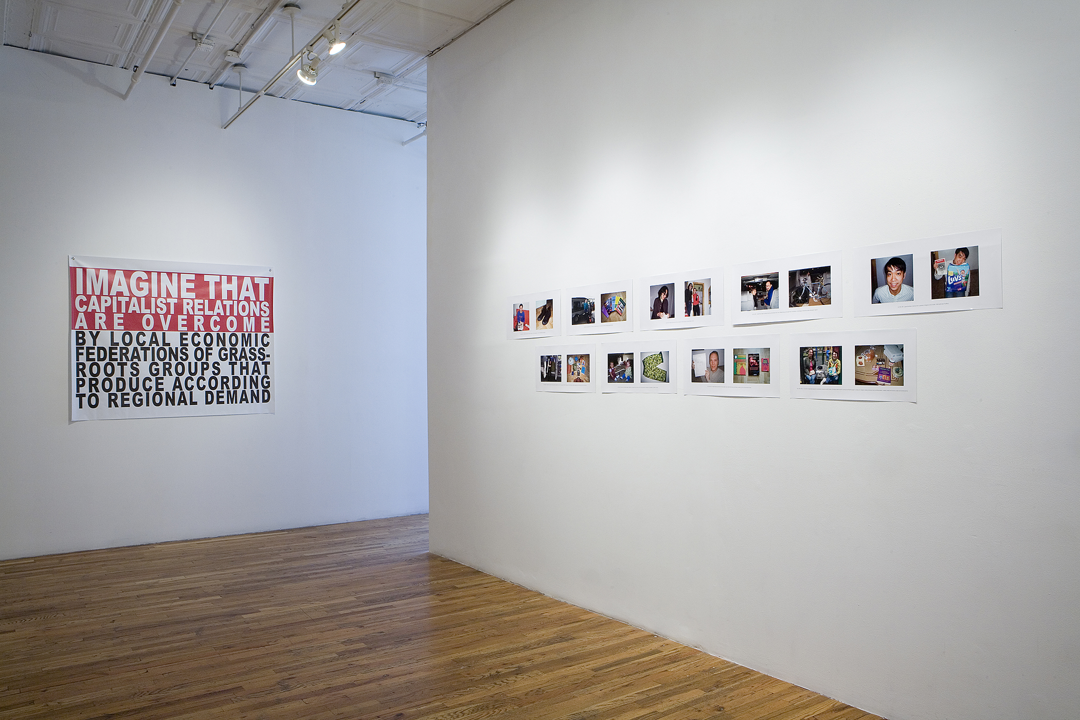 An installation view of two rows of photographic portraits tacked to the walls. In the background is a red, white and black print that says 