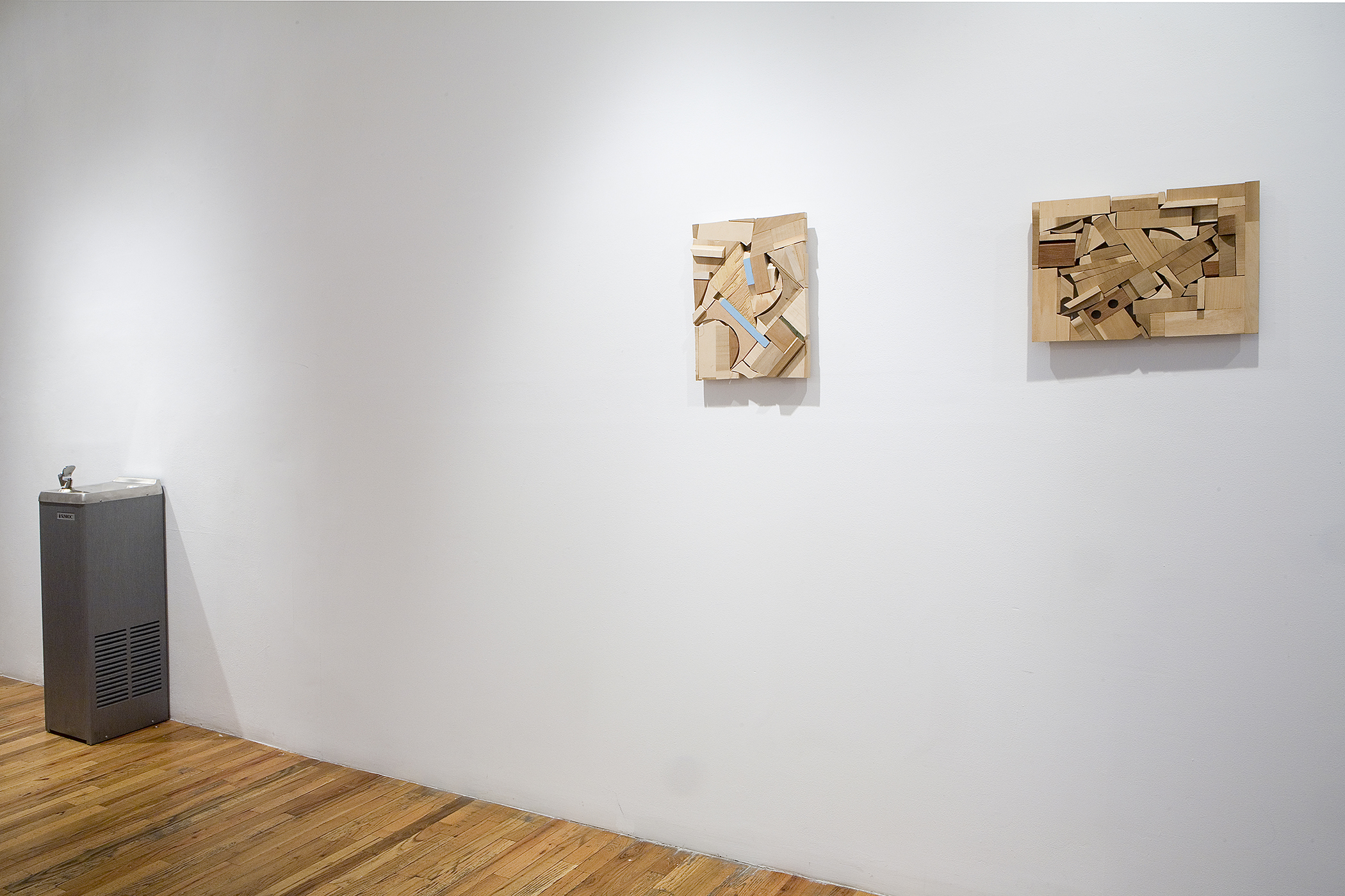 An installation view of two abstract wall works made of wood and a water fountain sculpture in the background.