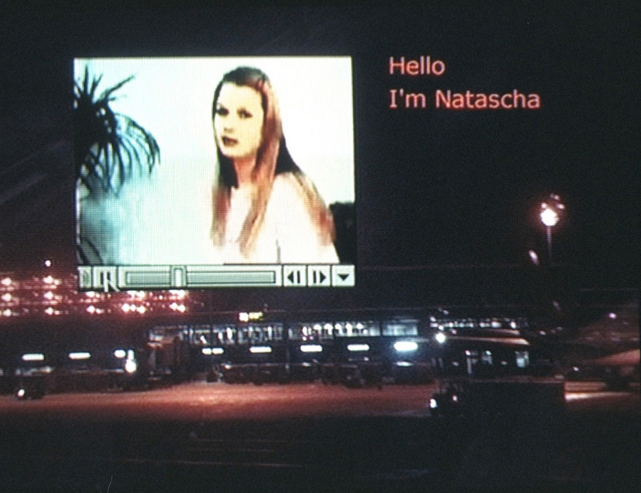 A nightime view of a truck driving past a parking lot. In the top left of the image is a superimposed video of a blond woman. The words 