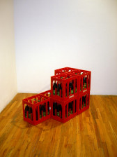 A corner installation view of red crates containing glass bottles of cola on a wood floor.