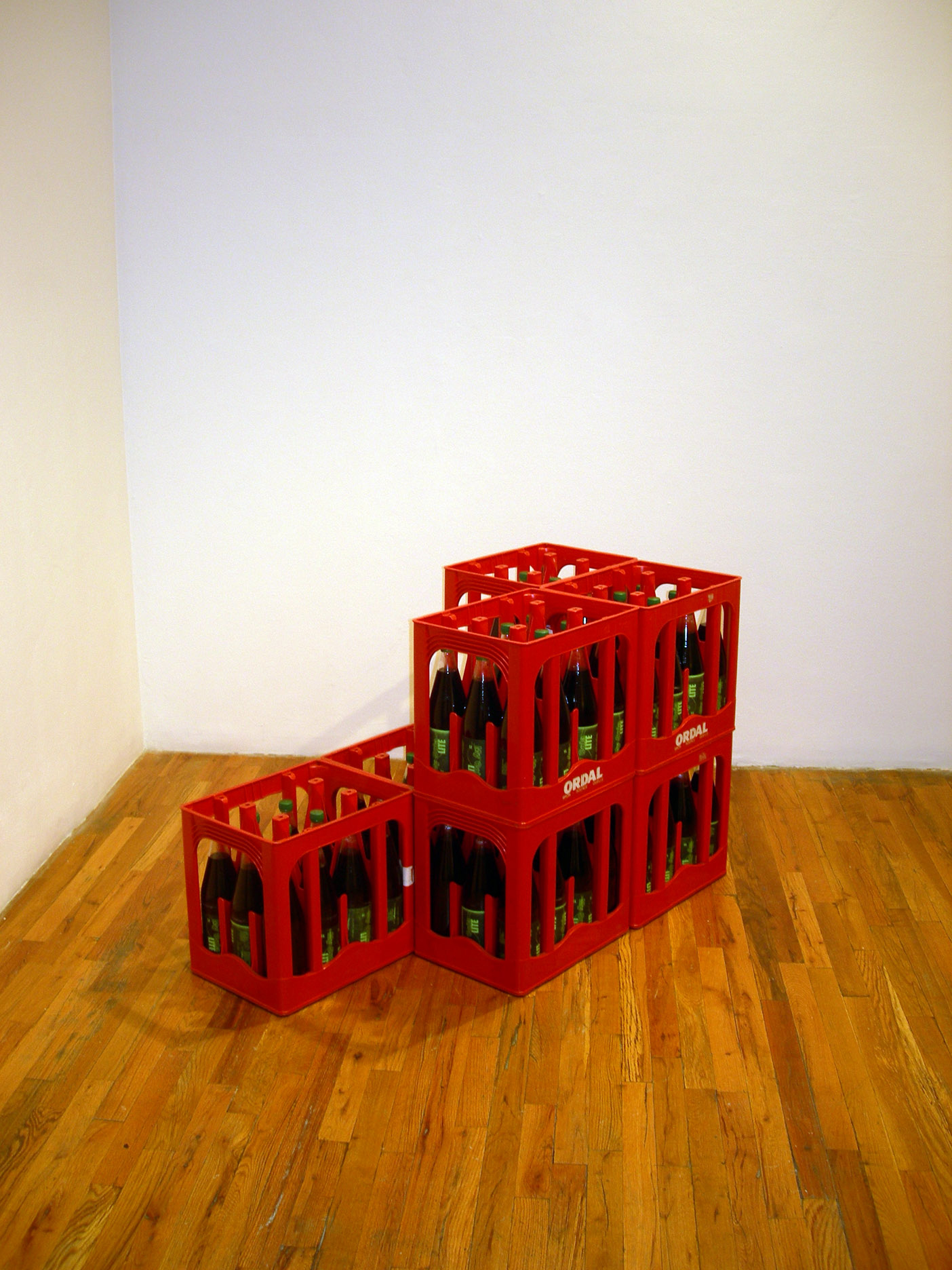 A corner installation view of red crates containing glass bottles of cola on a wood floor.