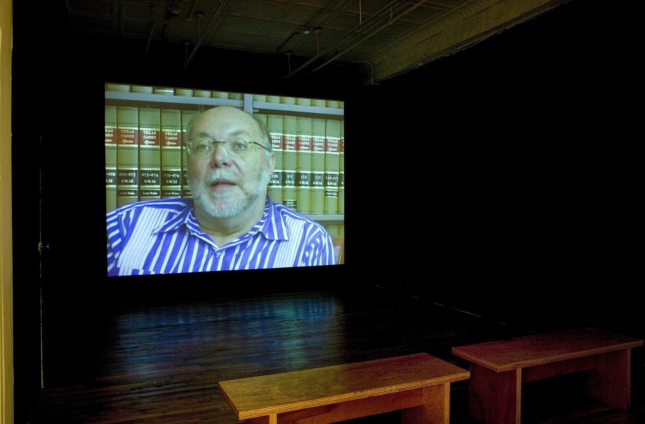 A black box screening room featuring a a video of a bespecaled white haired man with glasses speaking to the camera in front of rows of books.