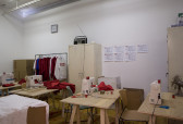 An installation view of multiple sewing stations and a clothing rack of red and white sweatshirts.