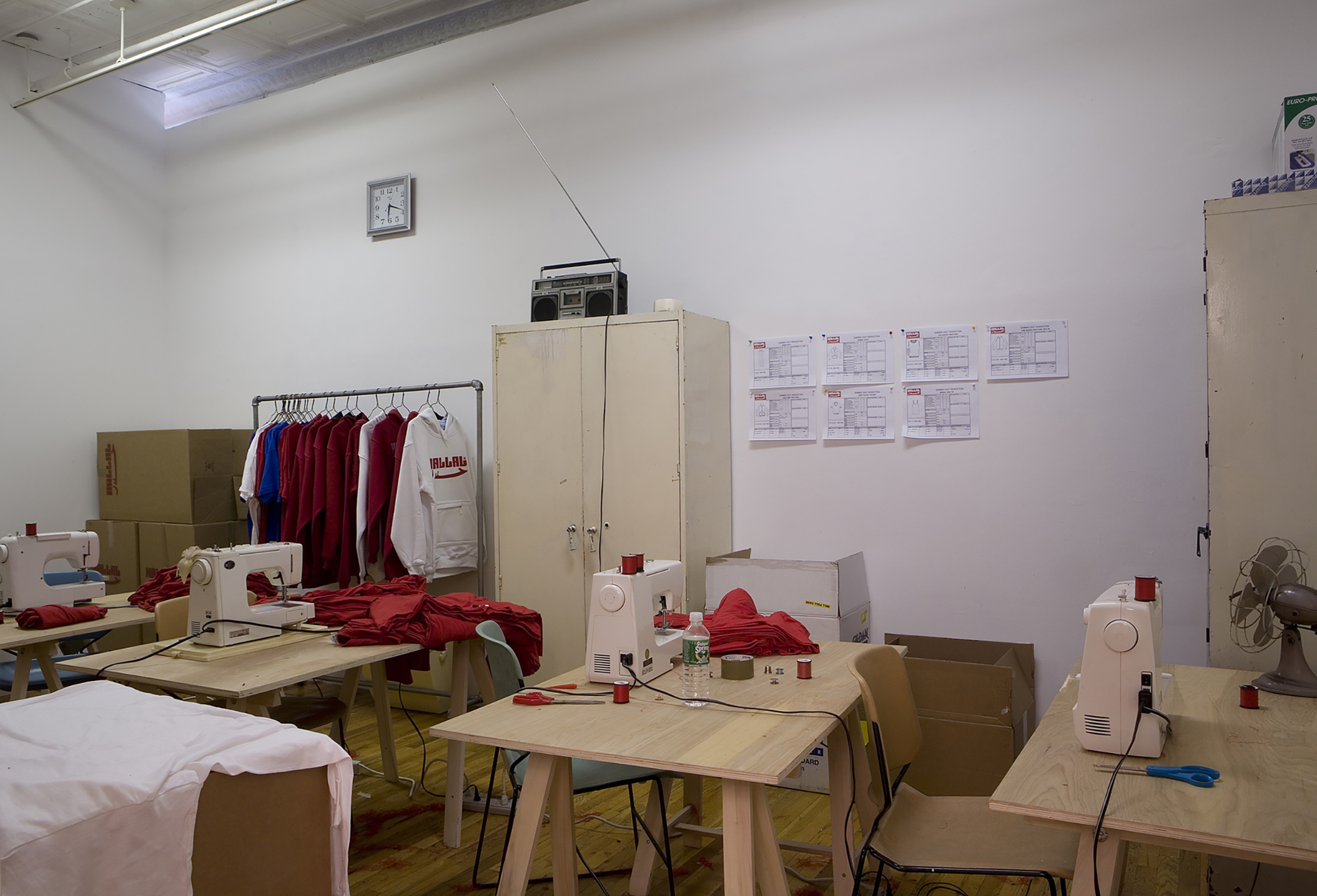 An installation view of multiple sewing stations and a clothing rack of red and white sweatshirts.