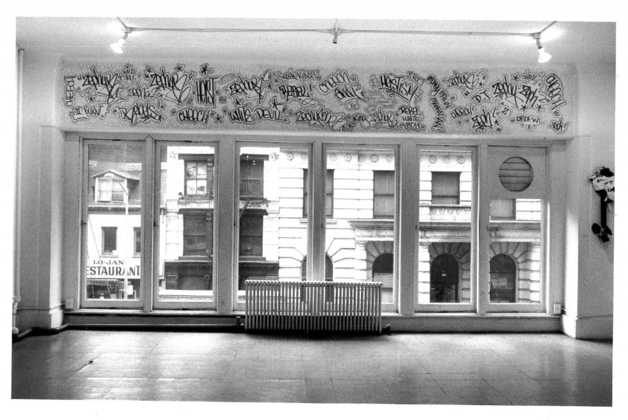 A view of a large window with the facade of other buildings, a restaurant is visible on the left. Graffiti text is visible above the length of the horizontal window and lights appear hanging from the ceiling directed to opposite sides.