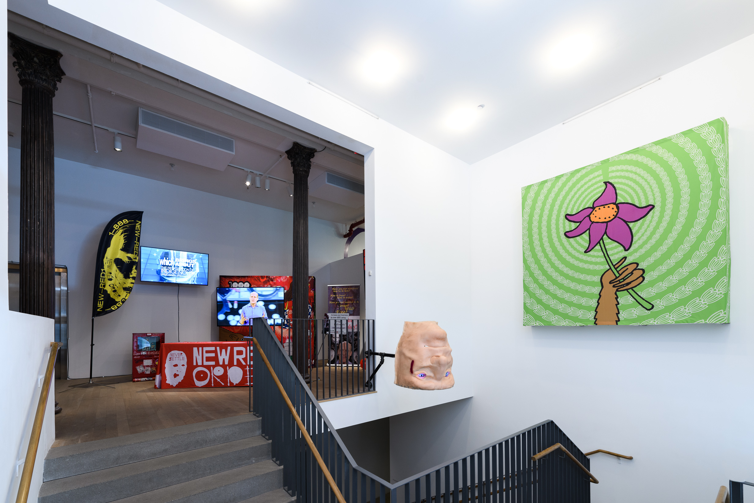 An image of an exhibition space with a sculpted, larger than life head. On the right is a large painting of a hand holding a flower, and on the left, is a table with red cover that reads 