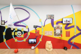 A multicolored wall with a collage of images, text, and tv monitors. A larger than life, sculpted head leans against the wall while beach balls scatter around it.