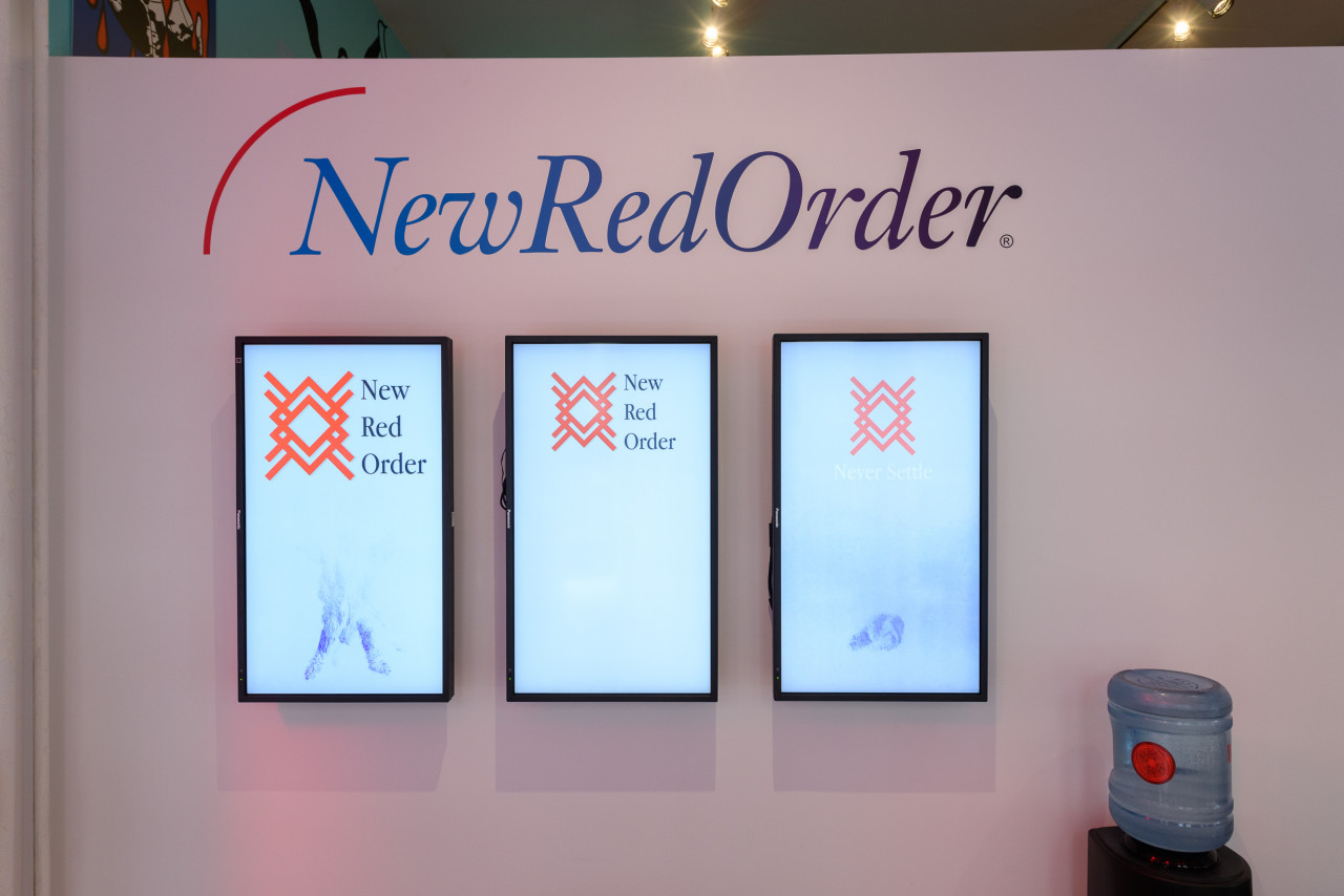 A close up image of three tv monitors on a wall with vinyl lettering that reads "New Red Order", in the right corner is a water cooler.